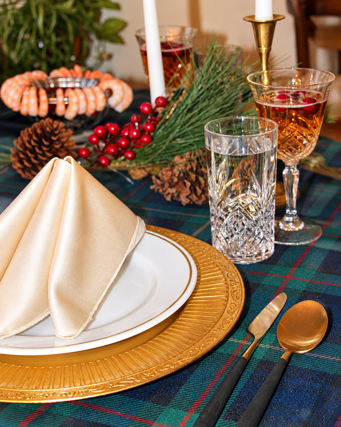 Christmas table setting with green and navy tartan linen, gold chargers, crystal glassware, ivory napkins and evergreen greenery.